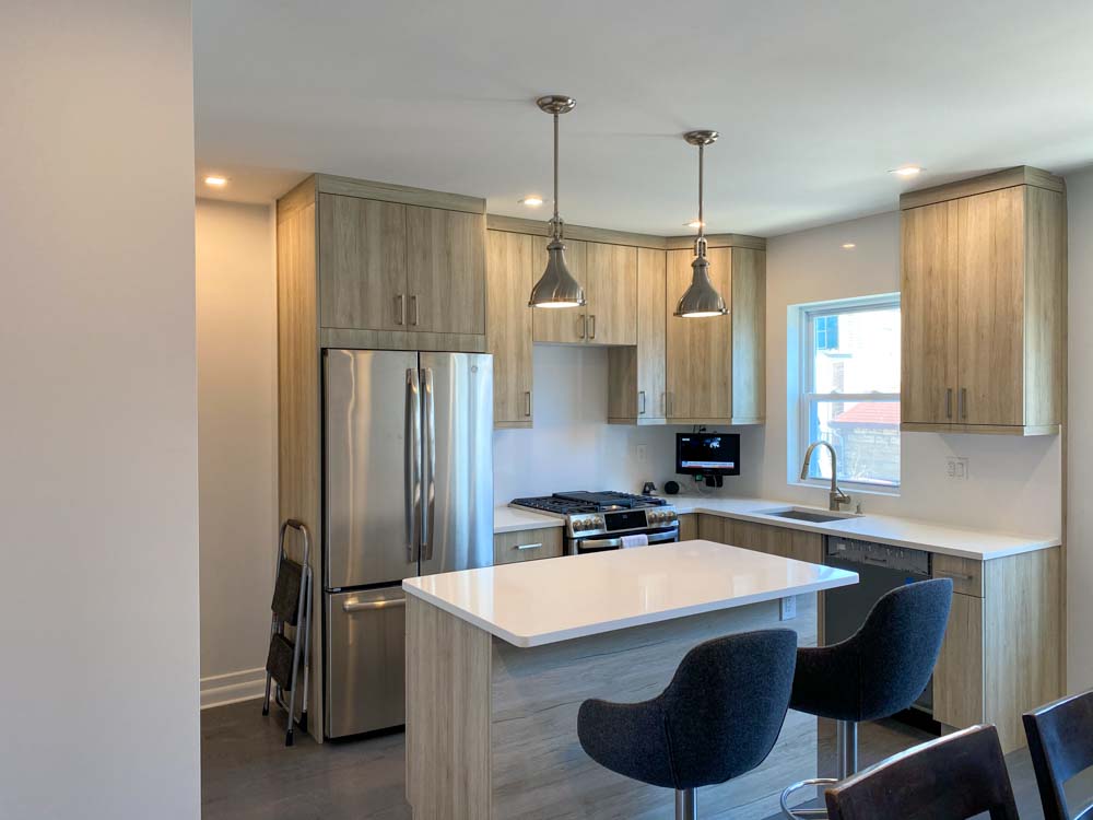 An apartment kitchen with stove range, refrigerator and cabinets around it, and an island bar with two high cushioned chair