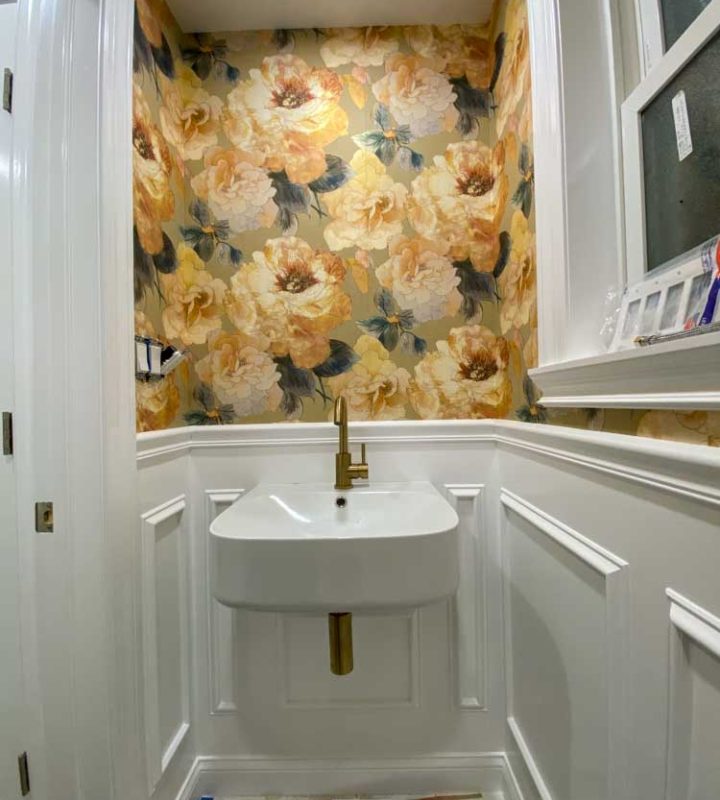 A powder room with floral designed wallpaper, a hanging sink with brass faucet